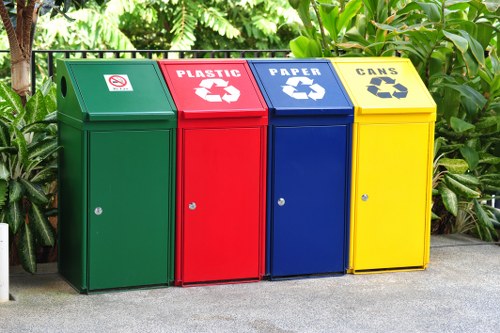 The image shows four distinct waste recycling bins arranged in a row on a concrete surface, surrounded by lush green foliage. The first bin on the left is green with a small white symbol indicating general waste or non-recyclable rubbish. Next is a red bin marked for plastic waste with a white recycling symbol and the word 'PLASTIC' printed above. The third bin is blue, designated for paper recycling, featuring a white recycling symbol and the word 'PAPER' printed on it. The fourth bin is yellow, intended for cans, with a black recycling symbol and the word 'CANS' displayed. Each bin has a small circular lock or latch on the front and a angled lid designed for easy access. The setting appears to be outdoors, possibly on a residential or commercial premises within or near Luton, reflecting effective waste segregation in line with local rubbish management practices. The branding references 'Commercial Waste Luton,' indicating a professional service specializing in waste disposal solutions in the area.