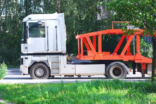 A large commercial flatbed truck parked on a paved road, with a white cab and an orange cargo area designed for transporting waste or construction materials. The truck's cab features a side mirror, windows, and black tires with metallic rims, while the open cargo bed is surrounded by a guardrail structure and is currently empty. The background includes green foliage and trees, suggesting a suburban or semi-rural environment near Luton, with sunlight illuminating the scene. The truck is positioned parallel to the grass verge and is part of a waste collection or removal operation, consistent with services provided by Commercial Waste Luton for rubbish disposal within the local area.