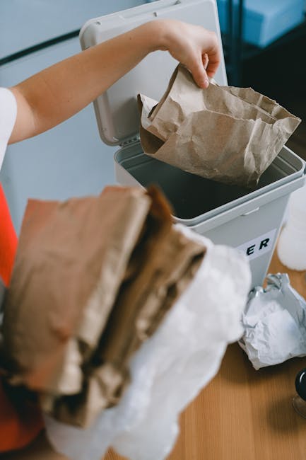 A person is placing a crumpled brown paper bag into a grey recycling bin, which is partially open and labeled for waste collection. The person’s arm is visible, wearing a short-sleeved shirt, and th