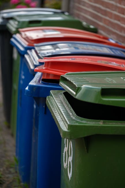 A person is placing a crumpled brown paper bag into a grey recycling bin, which is partially open and labeled for waste collection. The person’s arm is visible, wearing a short-sleeved shirt, and th